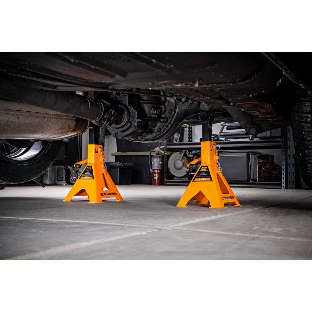 A pair of GEARWRENCH 3 ton ratcheting jack stands securely supporting the rear axle of a vehicle on a concrete garage floor, demonstrating professional shop application and stability.