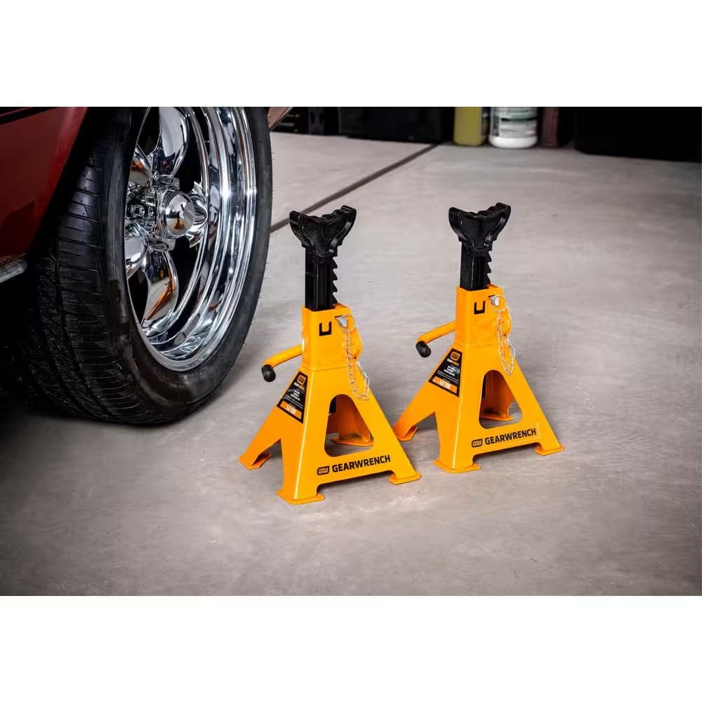 Two GEARWRENCH molten orange and black 3 ton ratcheting jack stands positioned on a level shop floor next to a vehicle tire, showing the high-visibility finish and wide base design.