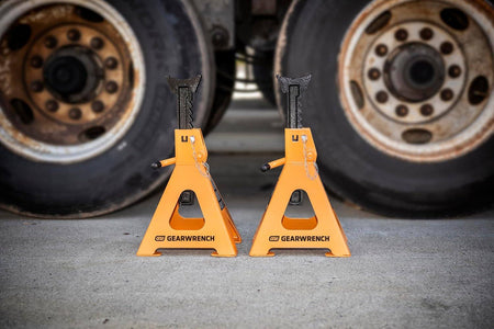 A pair of 6 ton GEARWRENCH jack stands positioned on a concrete shop floor in front of heavy-duty truck tires to demonstrate professional industrial application and stability.