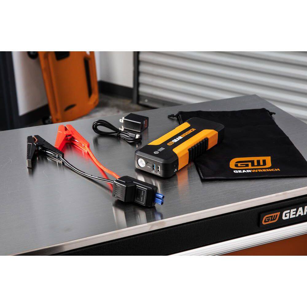 A complete product kit view of the GEARWRENCH GWLBJ20 set laid out on a workshop table. The image includes the 2500A jump starter unit, intelligent jumper cables, a USB wall charger, a charging cable, and a black drawstring storage bag with the GEARWRENCH logo.