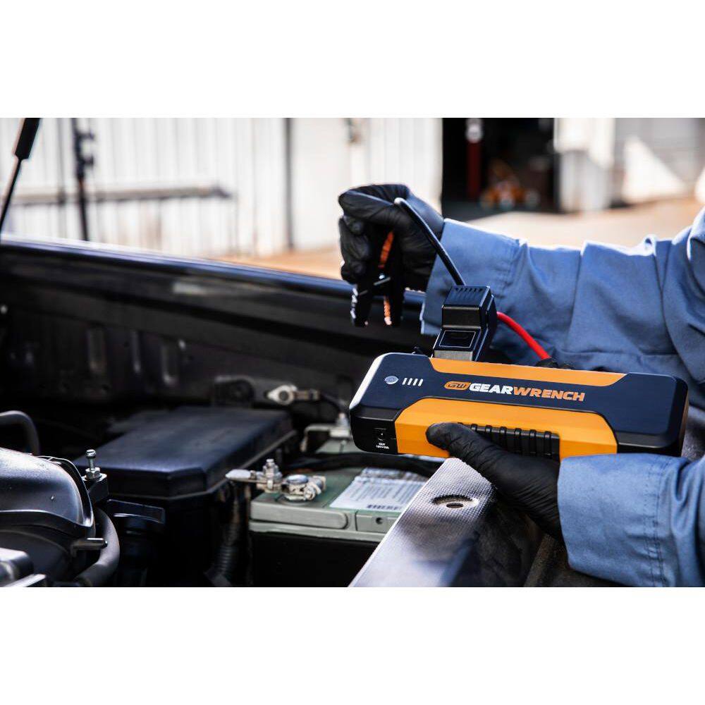 An action lifestyle shot of a technician using the GEARWRENCH 12V jump starter under the hood of a car. The professional, wearing black safety gloves, is shown connecting the intelligent cables to the vehicle's battery terminals to demonstrate the unit's portability.