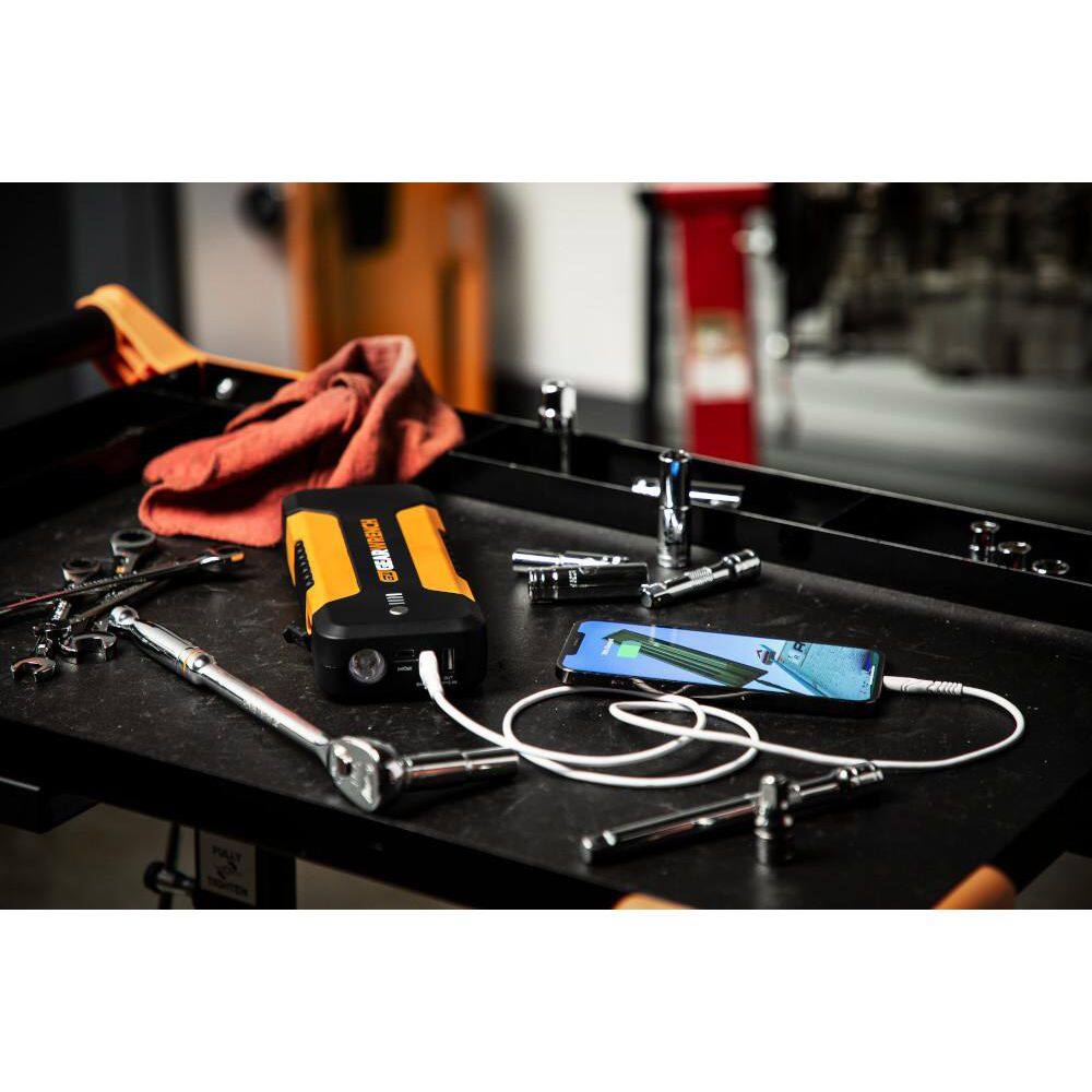 A lifestyle shot showing the GEARWRENCH jump starter being used as a power bank in a garage setting. The unit is resting on a mechanic's tool cart and is connected via a USB cable to charge a smartphone, surrounded by professional wrenches and tools.