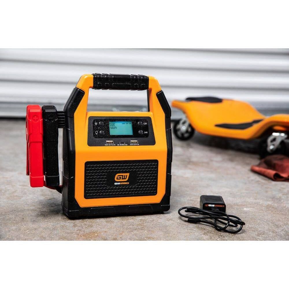 A product kit view showing the GEARWRENCH 12V/24V jump starter alongside its wall charging adapter and USB cable. The unit is shown in a garage setting, emphasizing its readiness as a professional-grade automotive battery maintainer and emergency power source.