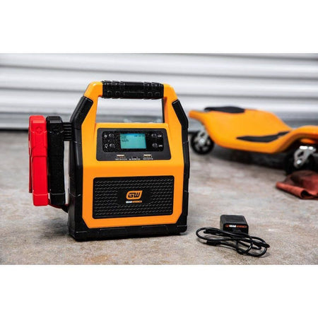 A product kit view showing the GEARWRENCH 12V/24V jump starter alongside its wall charging adapter and USB cable. The unit is shown in a garage setting, emphasizing its readiness as a professional-grade automotive battery maintainer and emergency power source.