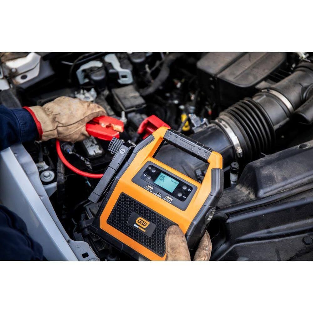 An action shot of a professional technician in protective gloves using the GEARWRENCH lithium jump starter. The worker is shown connecting the red clamp to a car battery under the hood, demonstrating the tool's portability and ease of use in a shop environment.