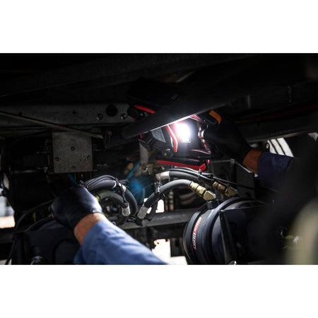 A lifestyle shot demonstrating the integrated 300-lumen LED flashlight on the GEARWRENCH jump starter. A worker in black gloves uses the bright light to illuminate the undercarriage and air brake system of a commercial vehicle during a nighttime inspection.