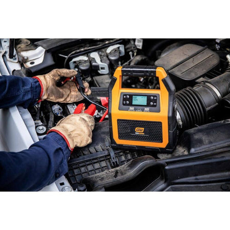 An action lifestyle shot of a technician using the GWLBJ30 unit to jump-start a 12V vehicle engine. The portable battery pack is shown resting on the engine shroud while the professional secures the heavy-duty clamps to the battery posts.