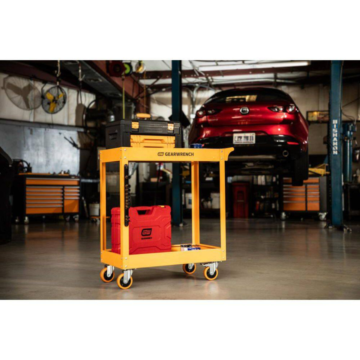 A lifestyle action shot of the GEARWRENCH molten orange service cart in a professional automotive garage. The cart is loaded with professional tool cases and a socket set, positioned next to a vehicle on a lift, demonstrating its utility and mobility in a real-world shop environment.