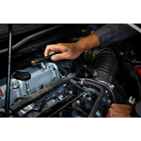 An action shot of the GEARWRENCH GWPL pen light being used to inspect a vehicle engine bay, with the magnetic clip attached to the underside of the hood for hands-free professional illumination.