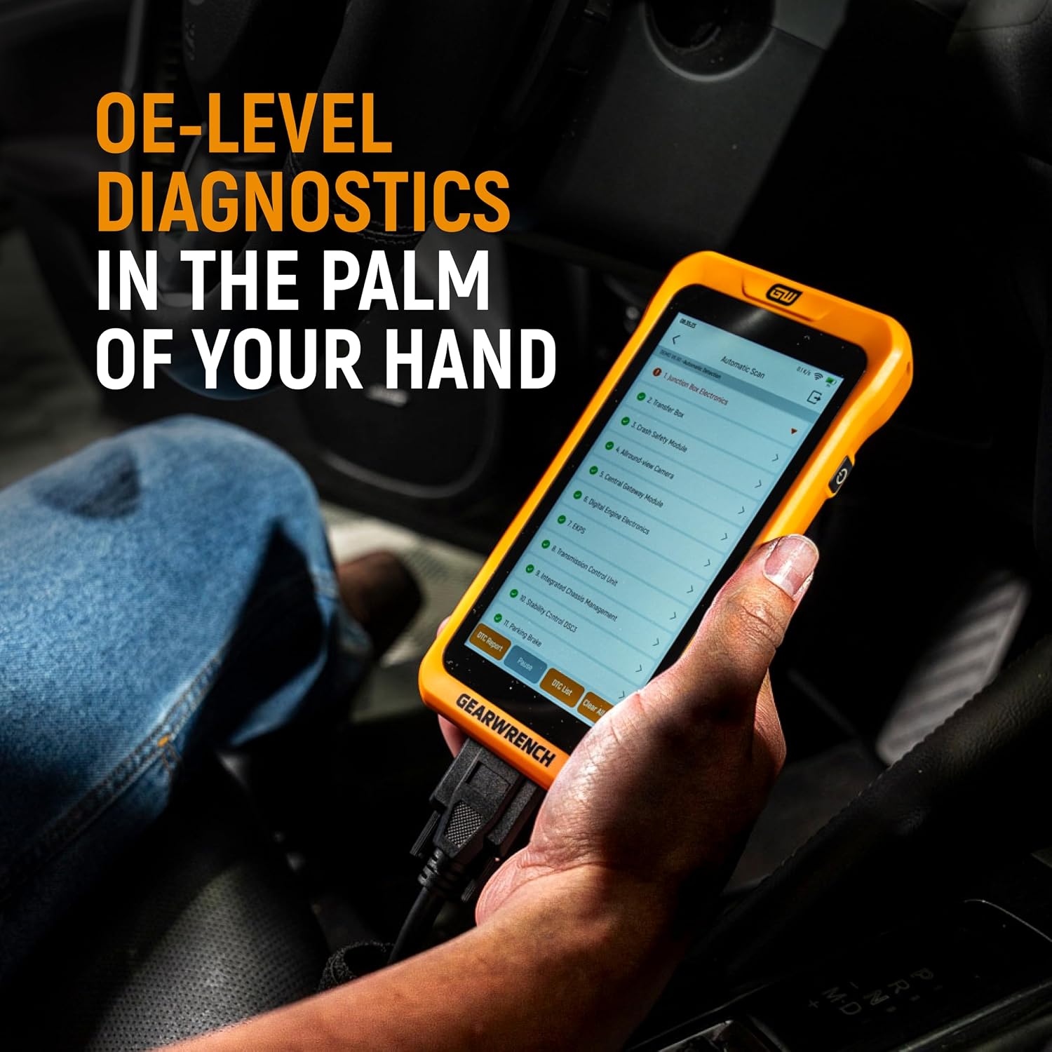 A close-up view of the GEARWRENCH electronic vehicle scan tool held by an auto mechanic inside a driver-side dashboard cockpit area running an active multi-system computer module sweep with orange frame trim visible.