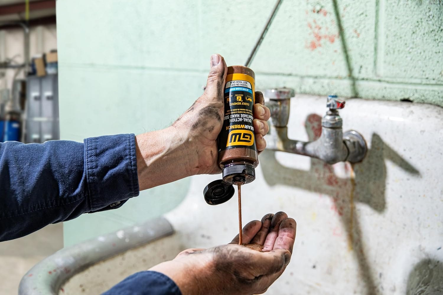 A technician with stained hands squeezing brown clay and pumice formula from an 8 ounce bottle of GEARWRENCH heavy duty hand cleaner. The solvent-free soap paste is dispensed at a commercial shop wash station sink to safely remove automotive grease, carbon buildup, and industrial grime from skin.