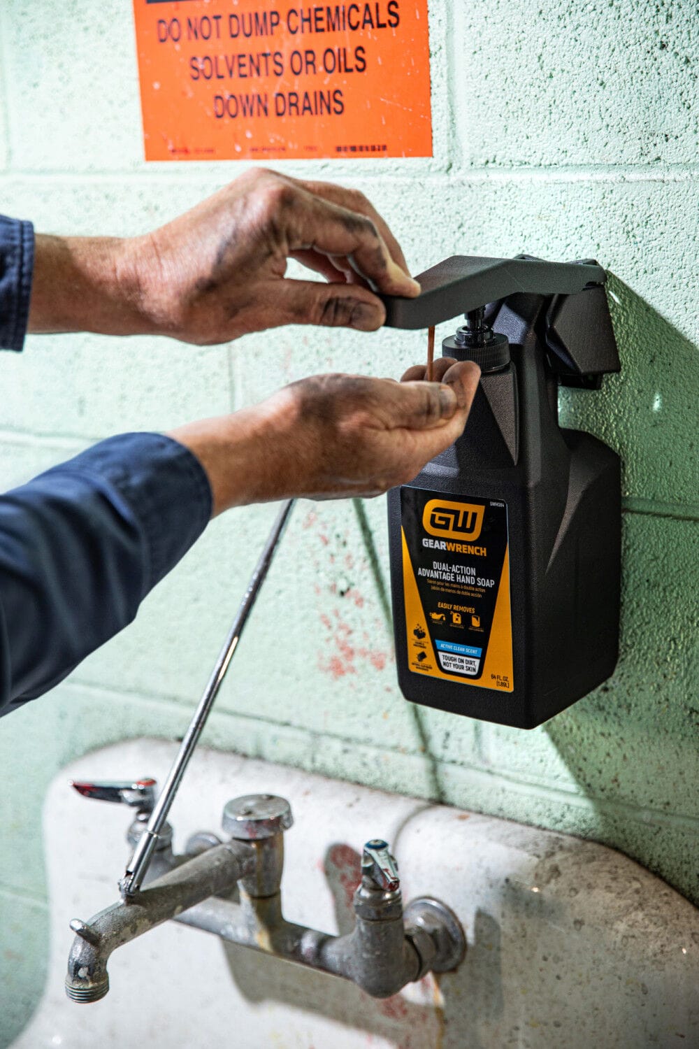 A worker with greasy hands pressing down on the black pump handle lever of a wall mounted GEARWRENCH GWDHS 64 ounce hand soap dispenser station. The structural bracket is anchored firmly to a green block utility wall above a commercial shop sink area, providing immediate mechanical hand cleaner access inside a busy industrial automotive workspace.