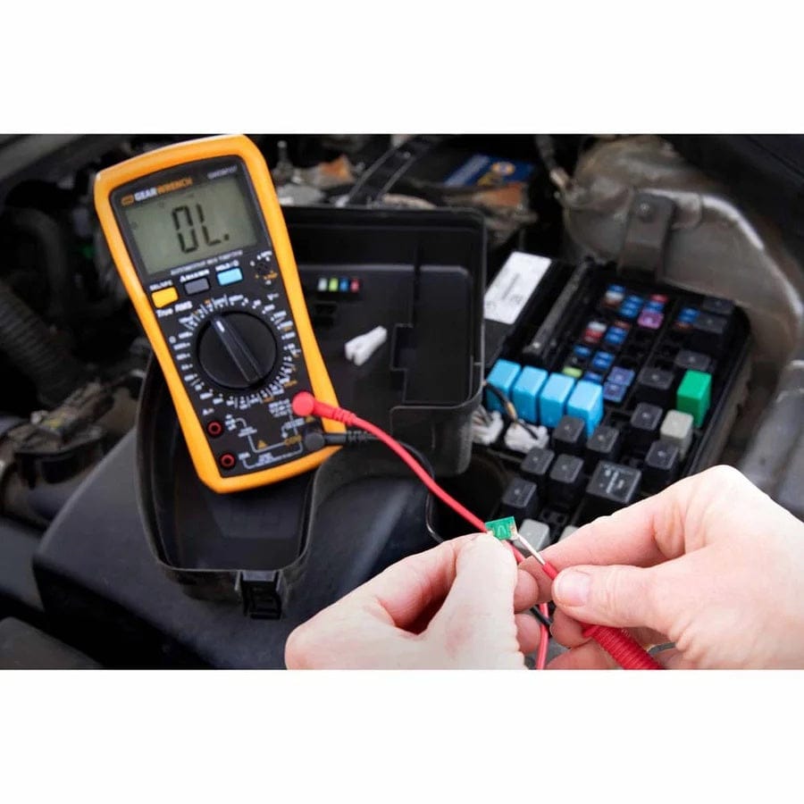 A mechanic's hands using the red and black test leads of the GEARWRENCH GWDM107 automotive digital multimeter to test a small green fuse near an open vehicle engine fuse box.