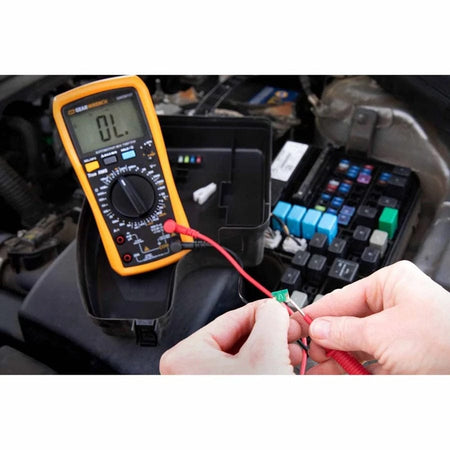 A mechanic's hands using the red and black test leads of the GEARWRENCH GWDM107 automotive digital multimeter to test a small green fuse near an open vehicle engine fuse box.