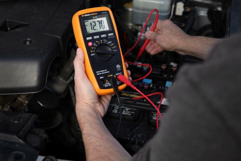 Technician using a GEARWRENCH GWDM015 digital multimeter to measure car battery voltage, with probes connected to terminals and display reading 12.70 volts.
