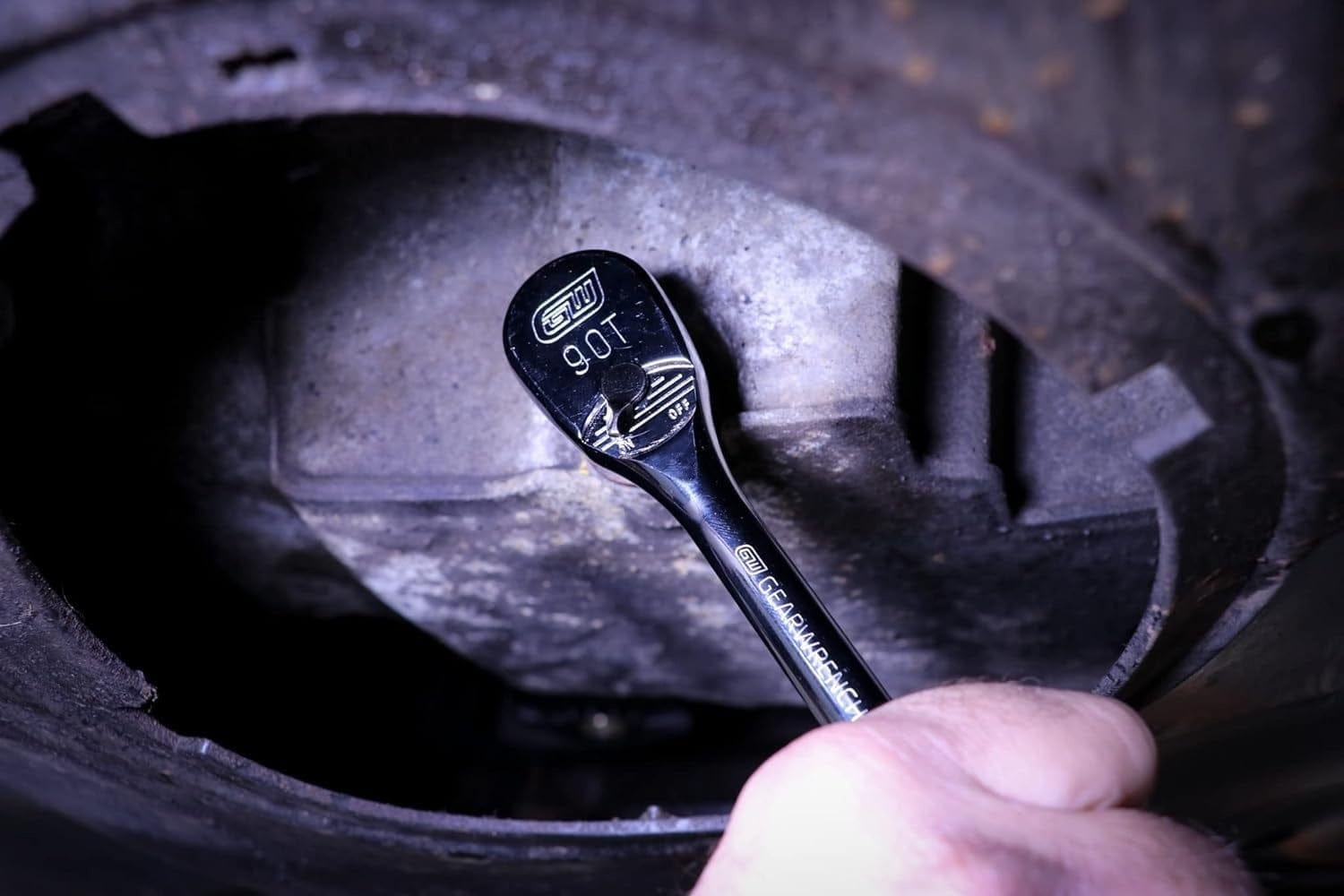 A hand uses a GearWrench 90-tooth ratchet in a confined space to turn a bolt on an engine block, demonstrating its tight swing arc.