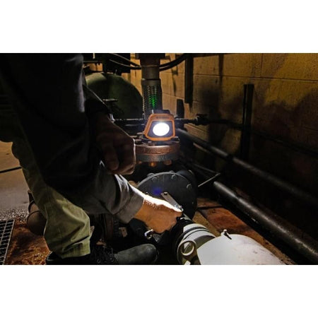 Person performing maintenance on industrial piping in a dimly lit room using a GearWrench LED shop light placed on a pipe fitting to illuminate the workspace