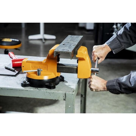 Person operating a bright yellow GearWrench bench vise mounted on a metal workbench, tightening a metal object between the jaws.