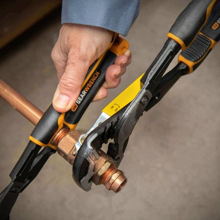 Close-up of a person using two GearWrench adjustable pliers to tighten a brass compression fitting on a copper pipe. The image highlights tool control and grip strength during plumbing installation.