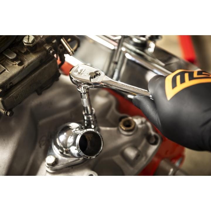 A close-up action shot of a gloved hand using a GEARWRENCH ratchet and extension to work on a car engine's thermostat housing.
