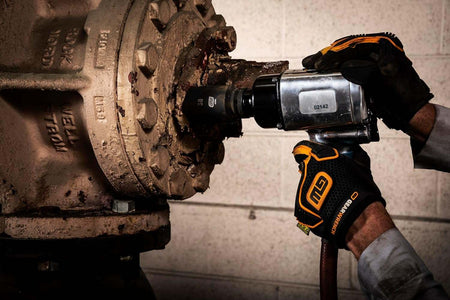 A close-up action shot of a gloved hand using a 3/4-inch air impact gun and a GEARWRENCH socket to remove a large, rusty bolt from an industrial pipe flange.