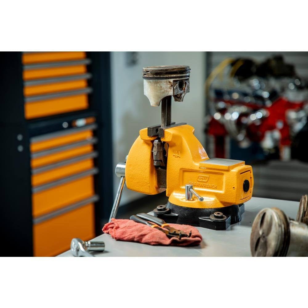 Bright yellow bench vise mounted on a workbench holding a worn piston, with tools and engine parts visible in a mechanical repair workspace.