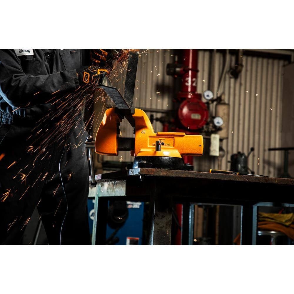 Worker using a power tool to grind metal clamped in a bright orange bench vise mounted on a workbench, with sparks flying in an industrial workshop setting
