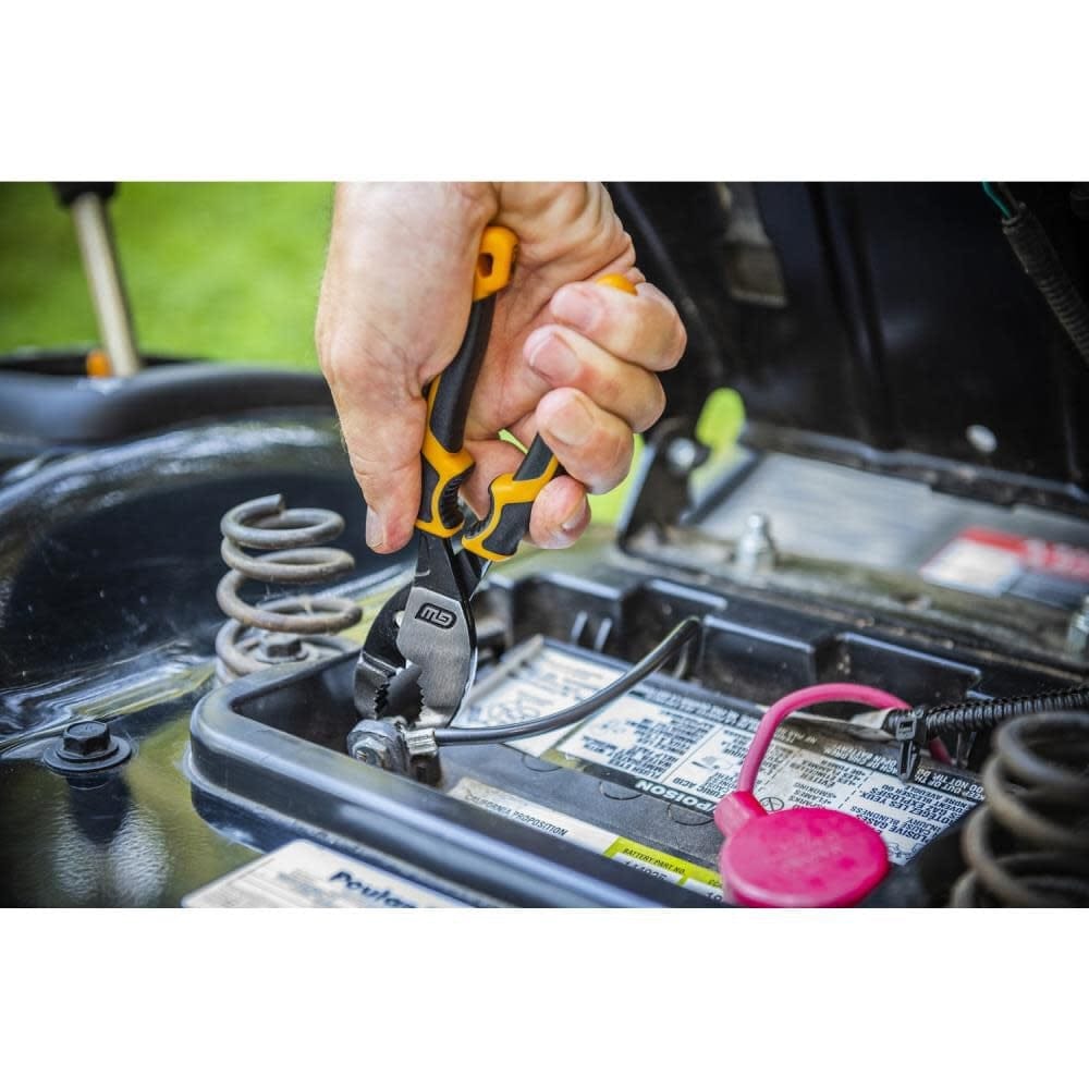 Person using black and yellow GearWrench pliers to tighten a bolt on a battery terminal installed in machinery; visible battery labels and surrounding mechanical parts.