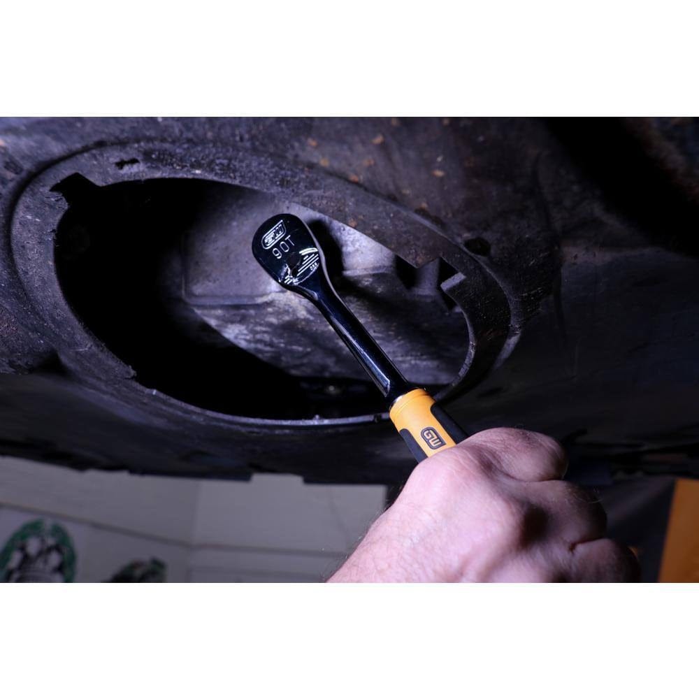Close-up of a hand using a ratchet wrench with yellow and black handle on a vehicle’s undercarriage in a workshop.