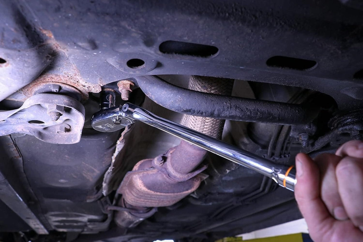 A close-up action shot of a hand using the GEARWRENCH flex-head ratchet to access a rusty bolt on the undercarriage of a car.