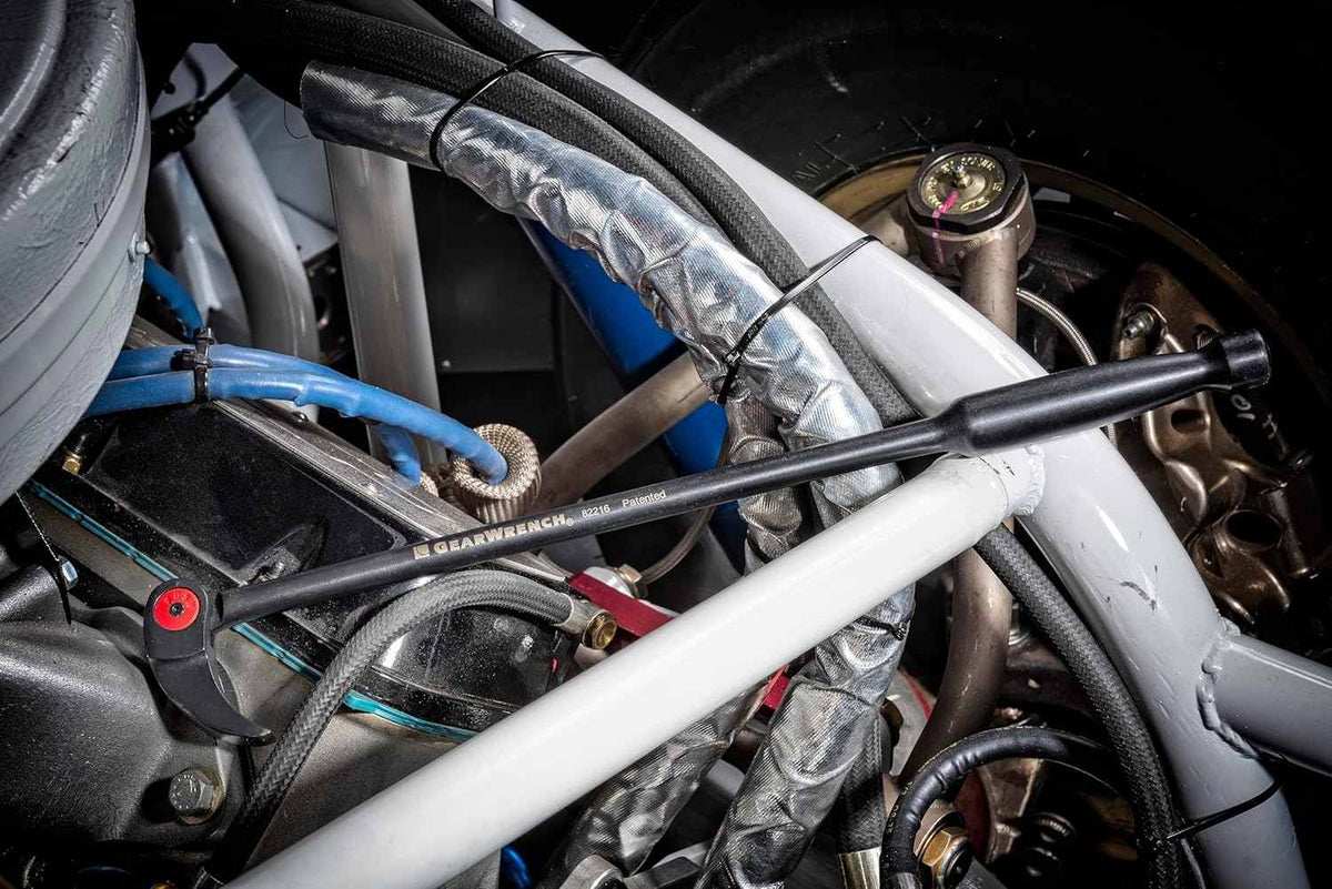 Lifestyle shot showing the pry bar reaching into a crowded engine bay with silver heat wrapping and blue hoses.