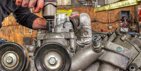 Person using a power tool on a mechanical engine component in a workshop, surrounded by pulleys and metal casings.