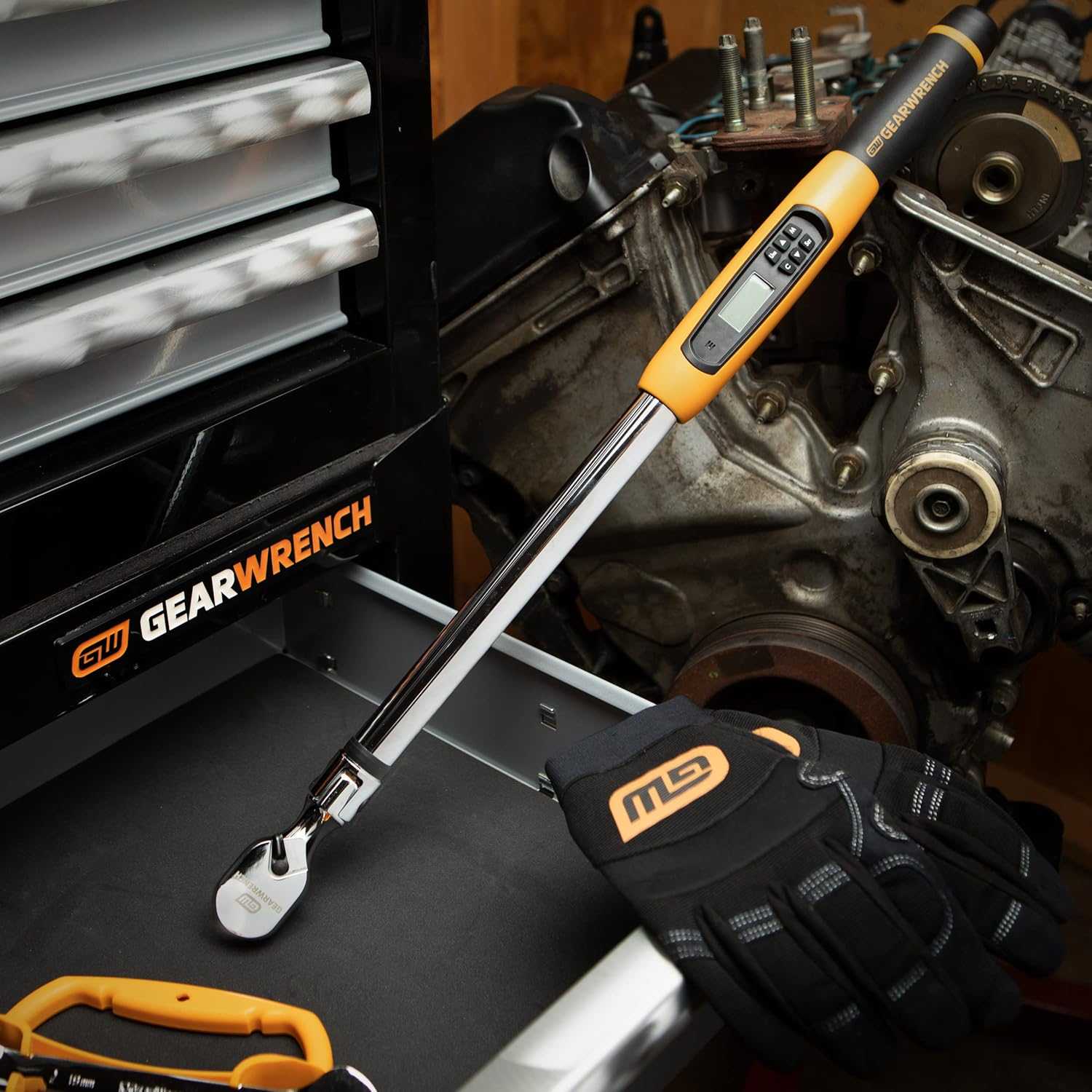 GearWrench digital torque wrench with display and buttons shown on a workbench beside gloves and engine block.