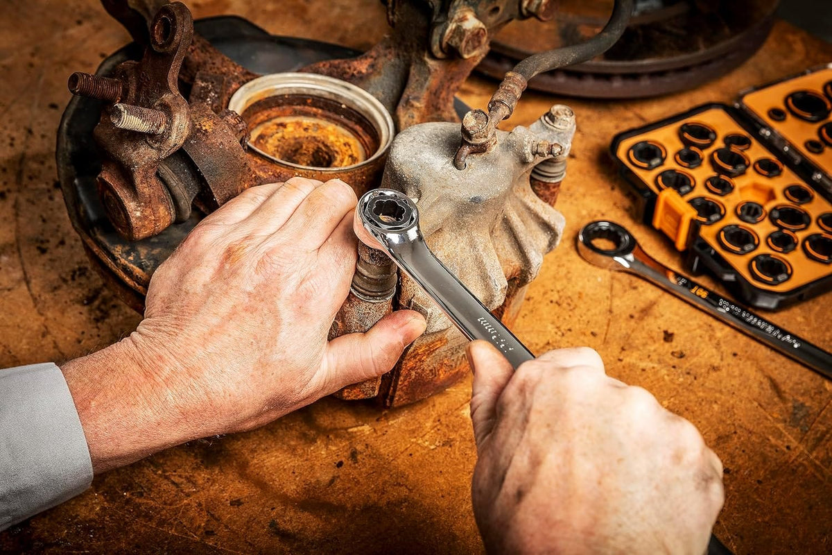 Action shot demonstrating a GearWrench Bolt Biter insert being used with a ratcheting wrench to remove a heavily rusted and damaged bolt from a vehicle's suspension component.