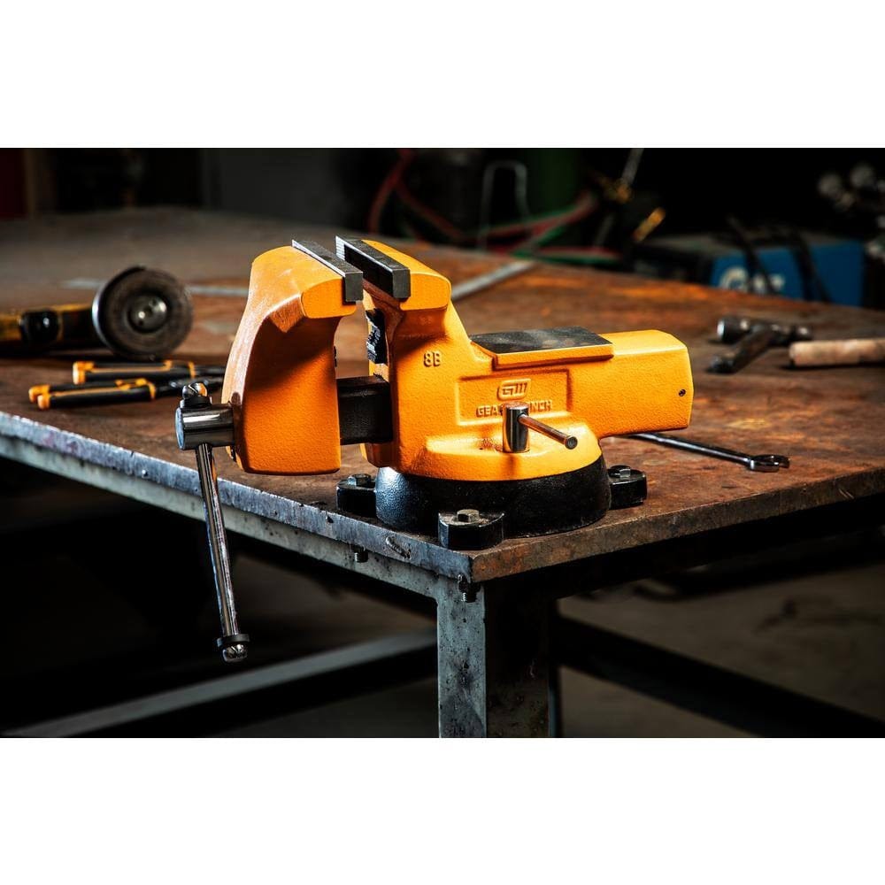A GearWrench 8 inch professional mechanic's bench vise mounted on a metal industrial workbench in a shop environment, showcasing its molten orange finish and heavy-duty anvil surface.