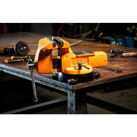 A GearWrench 8 inch professional mechanic's bench vise mounted on a metal industrial workbench in a shop environment, showcasing its molten orange finish and heavy-duty anvil surface.