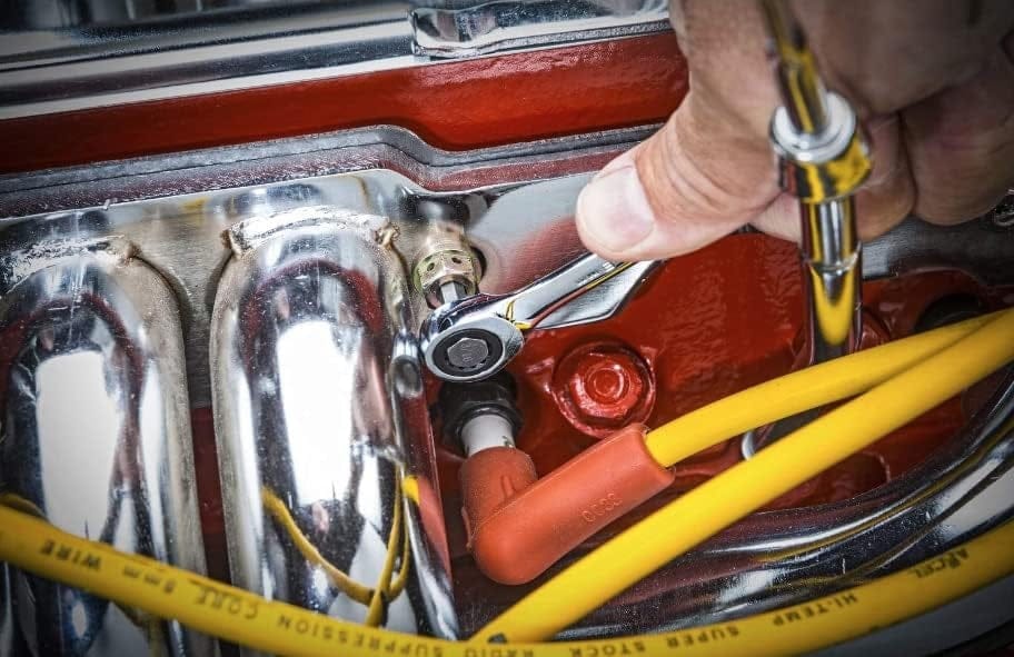 Close-up action shot of a mechanic using a small ratcheting wrench to adjust a fastener on a red engine valve cover near spark plug wires.