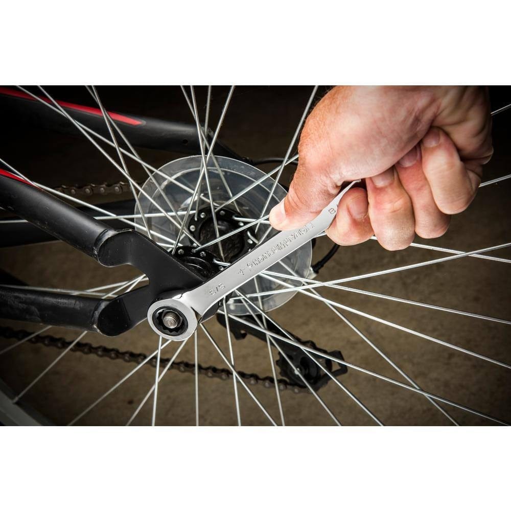 A close-up action shot of a hand using a GearWrench ratcheting wrench to tighten a bolt on a bicycle wheel. The image demonstrates the wrench's ability to operate in a restricted space between the bicycle's spokes.