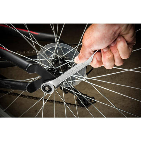 A close-up action shot of a hand using a GearWrench ratcheting wrench to tighten a bolt on a bicycle wheel. The image demonstrates the wrench's ability to operate in a restricted space between the bicycle's spokes.