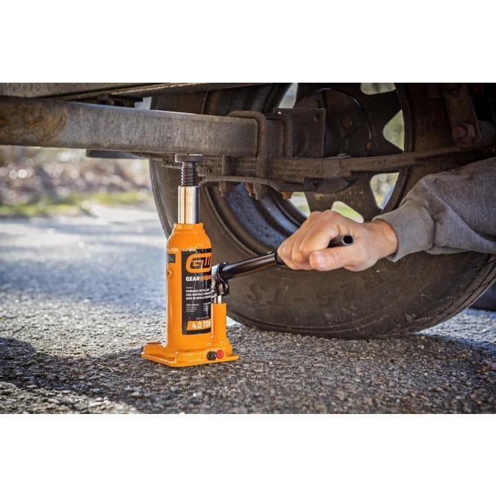 Person using Gearwrench 4 Ton hydraulic bottle jack to lift vehicle axle during repair; jack positioned under suspension.