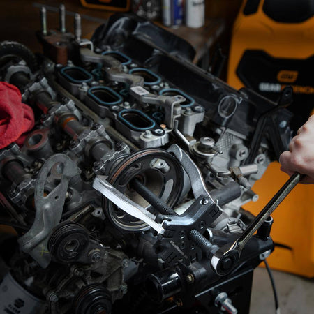 An automotive technician using the Gearwrench 5-ton ratcheting puller with a hand wrench to remove a pulley from a vehicle engine, demonstrating the tool's indexing leg design and one-handed setup capability.