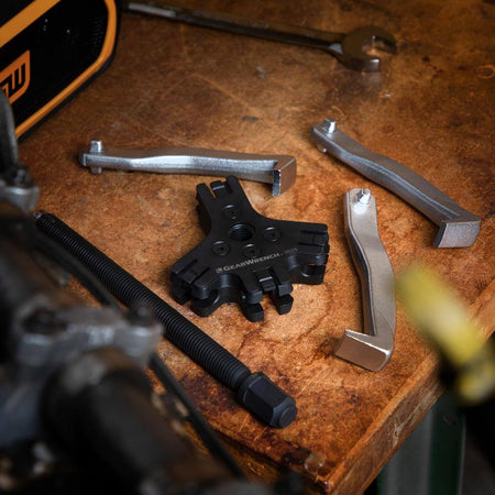 A professional Gearwrench indexing puller head and three forged steel reversible jaws laid out on a wooden workbench, demonstrating the modular components of the 3627 ratcheting puller set used for specialized automotive bearing and gear removal.