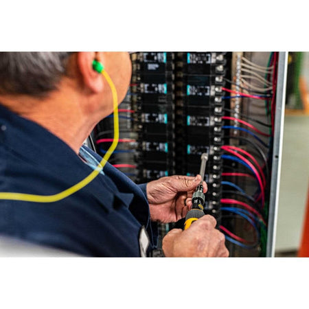 Close-up action shot of an electrician wearing safety earplugs using the Gearwrench torque screwdriver with a long nut driver bit to secure connections inside a high-voltage electrical breaker panel.