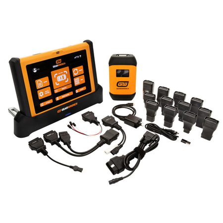 An overhead workspace display showcasing the complete Gearwrench GWSMART10 10-inch diagnostic scanner configuration, including the main touchscreen tablet seated in its black rapid-charge dock, the wireless vehicle communication interface VCI module, an assortment of vehicle-specific adapter plugs, and data connectivity cables laid out on a clean white surface.