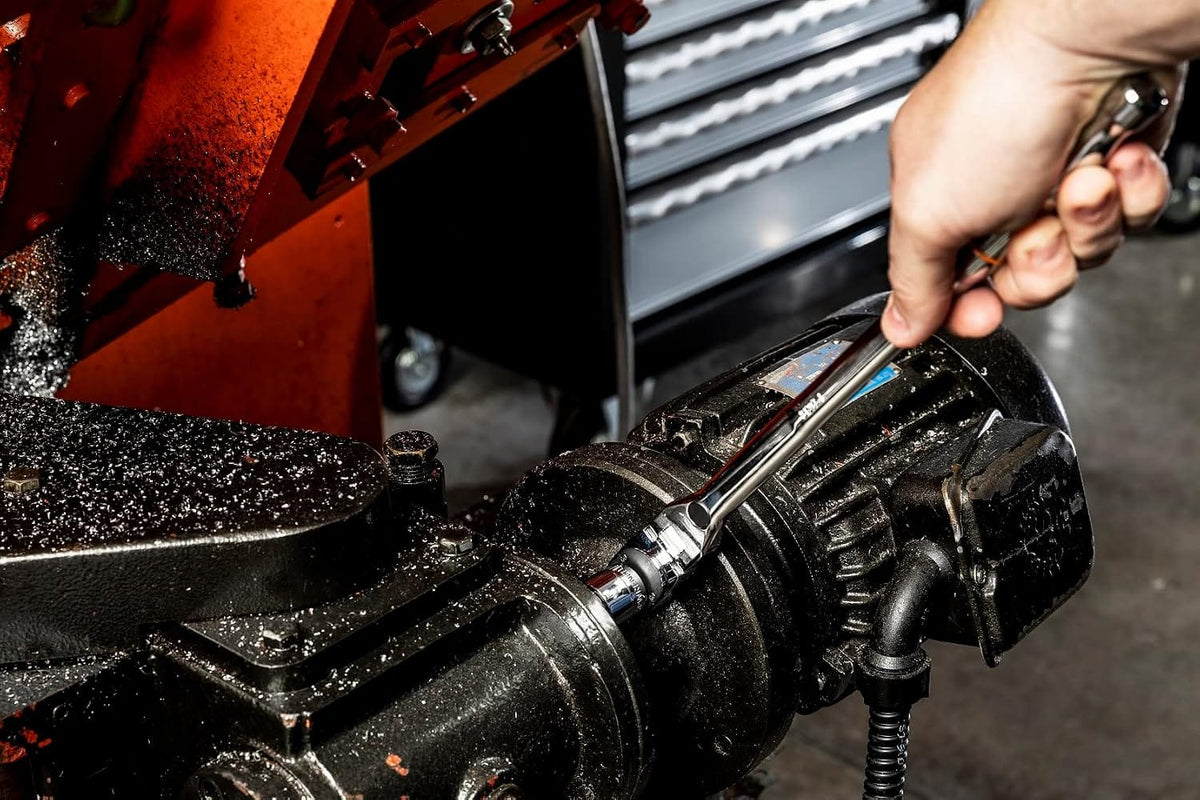 Close-up action shot of a technician using the Gearwrench 3/8-inch drive locking flex head ratchet and a stubby metric socket to loosen a bolt on a black industrial electric motor housing in a machine shop environment.
