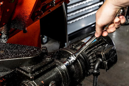 Close-up action shot of a technician using the Gearwrench 3/8-inch drive locking flex head ratchet and a stubby metric socket to loosen a bolt on a black industrial electric motor housing in a machine shop environment.