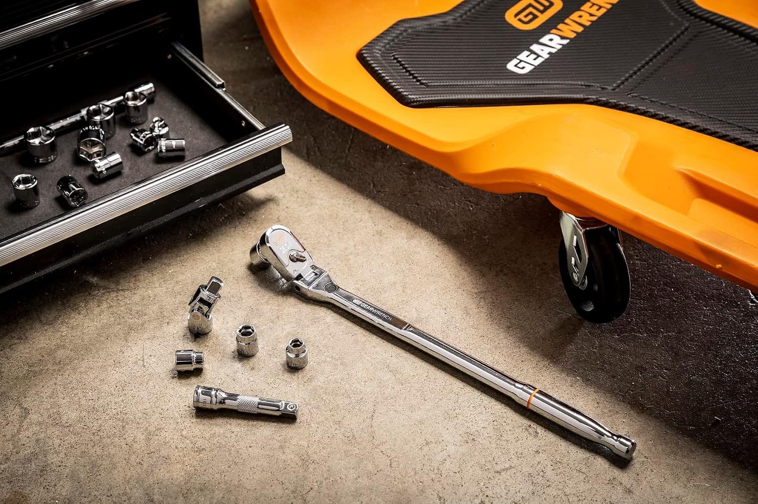 High-angle workshop shot showing the full polish chrome Gearwrench 120XP flex head ratchet resting on a concrete floor next to an orange mechanic creeper, with various stubby metric sockets, a universal joint, an extension bar, and an open tool chest drawer visible in the background.