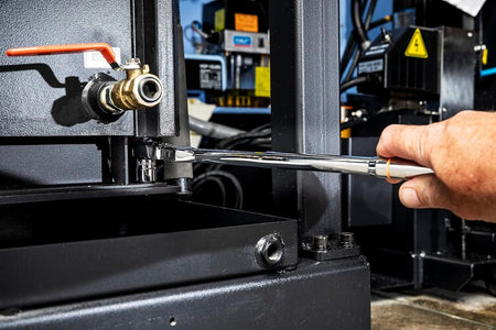 Close-up action shot of a person using a full polish chrome Gearwrench locking flex head ratchet and stubby SAE socket to reach and turn a low-clearance bolt underneath a valve assembly on black industrial plant machinery.