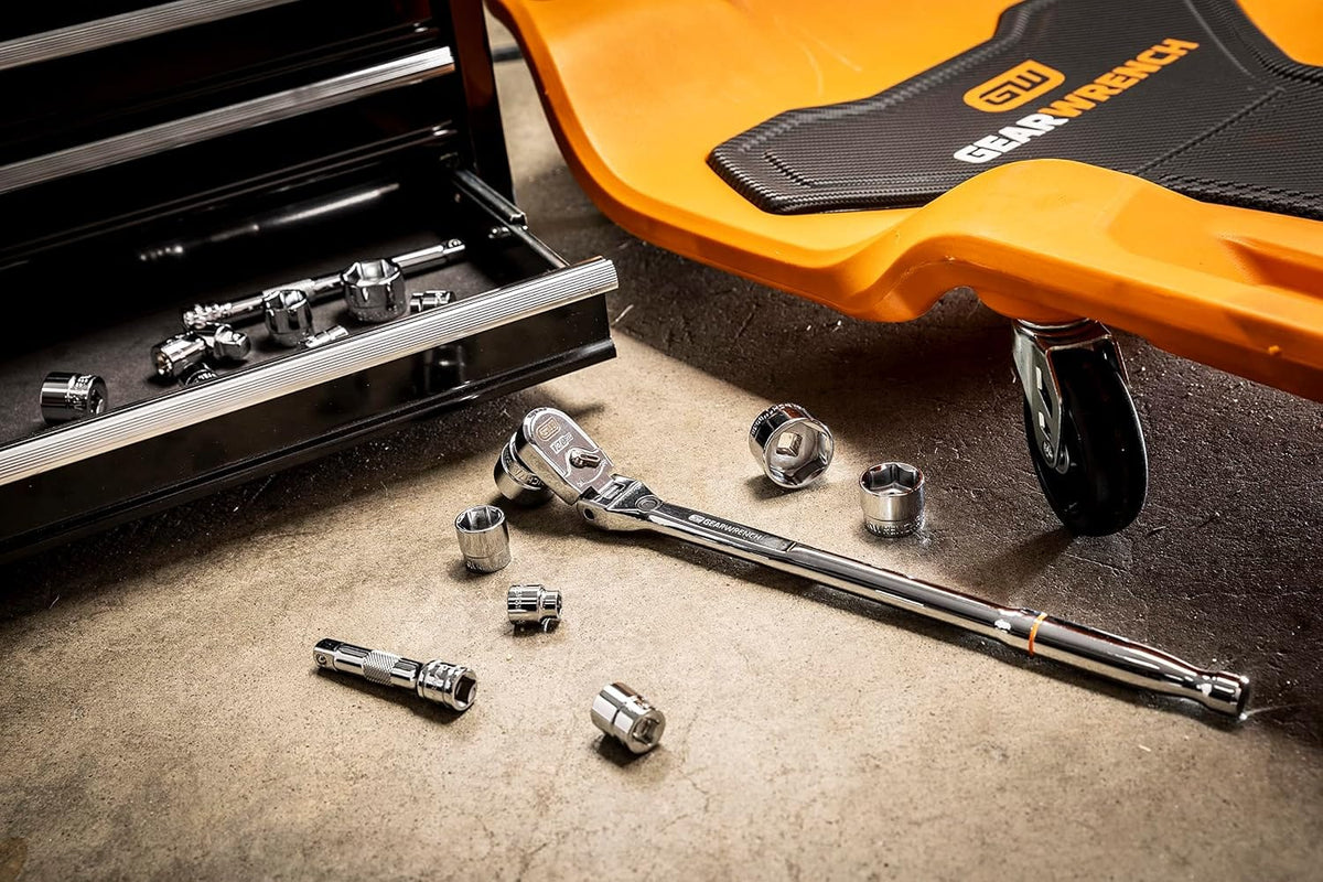 High-angle workshop floor photograph featuring the chrome Gearwrench 120XP flex head ratchet surrounded by loose stubby 6-point SAE sockets and an extension bar, situated next to an orange mechanic creeper and an open tool chest drawer.