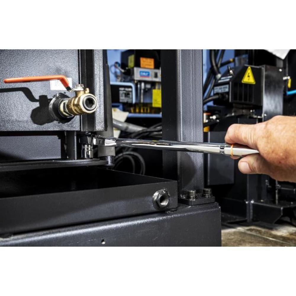 Horizontal wide-angle action view of a field mechanic applying leverage with a 3/8-inch drive Gearwrench flex head ratchet to complete a difficult clearance adjustment on heavy industrial manufacturing equipment.